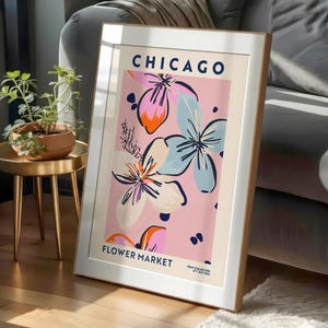May include: A framed vertical art print featuring the text CHICAGO FLOWER MARKET in dark blue. The design displays stylized floral illustrations in shades of pink, light blue, and orange against a pale pink background with dark blue accents.