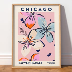 May include: A framed wall art print featuring the text CHICAGO FLOWER MARKET PRINT COLLECTION N&deg;1 JULY 2022. The design displays stylized floral illustrations in shades of pink, light blue, and orange against a soft pink background.