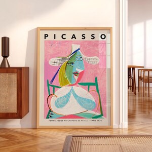 May include: A framed art print of Pablo Picasso's 1938 painting, Femme Assise au Chapeau de Paille. The artwork features a stylized figure in a hat against a pink background, with the text PICASSO at the top and the title at the bottom.