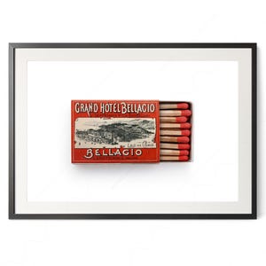 May include: A framed print featuring a vintage matchbox. The matchbox is red with the words "GRAND HOTEL BELLAGIO" and a detailed illustration of a lakeside town. Several wooden matches with red tips are visible.