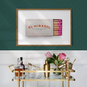 K&ouml;nnte beinhalten: Ein gerahmter Druck mit einer Streichholzschachtel mit dem Text "El Mirasol Una Cocina Mexicana" in Orange. Die Streichholzk&ouml;pfe sind rosa. Ein goldener Servierwagen mit Gl&auml;sern, einem Cocktailshaker und Blumen befindet sich im Vordergrund.