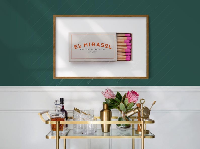 K&ouml;nnte beinhalten: Ein gerahmter Druck mit einer Streichholzschachtel mit dem Text "El Mirasol Una Cocina Mexicana" in Orange. Die Streichholzk&ouml;pfe sind rosa. Ein goldener Servierwagen mit Gl&auml;sern, einem Cocktailshaker und Blumen befindet sich im Vordergrund.