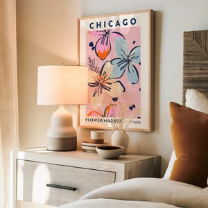 May include: A framed wall art print titled CHICAGO FLOWER MARKET with a pink background and abstract floral illustrations in blue, orange, and black. The print is displayed in a light wood frame above a bedside table with a lamp and ceramic bowls.