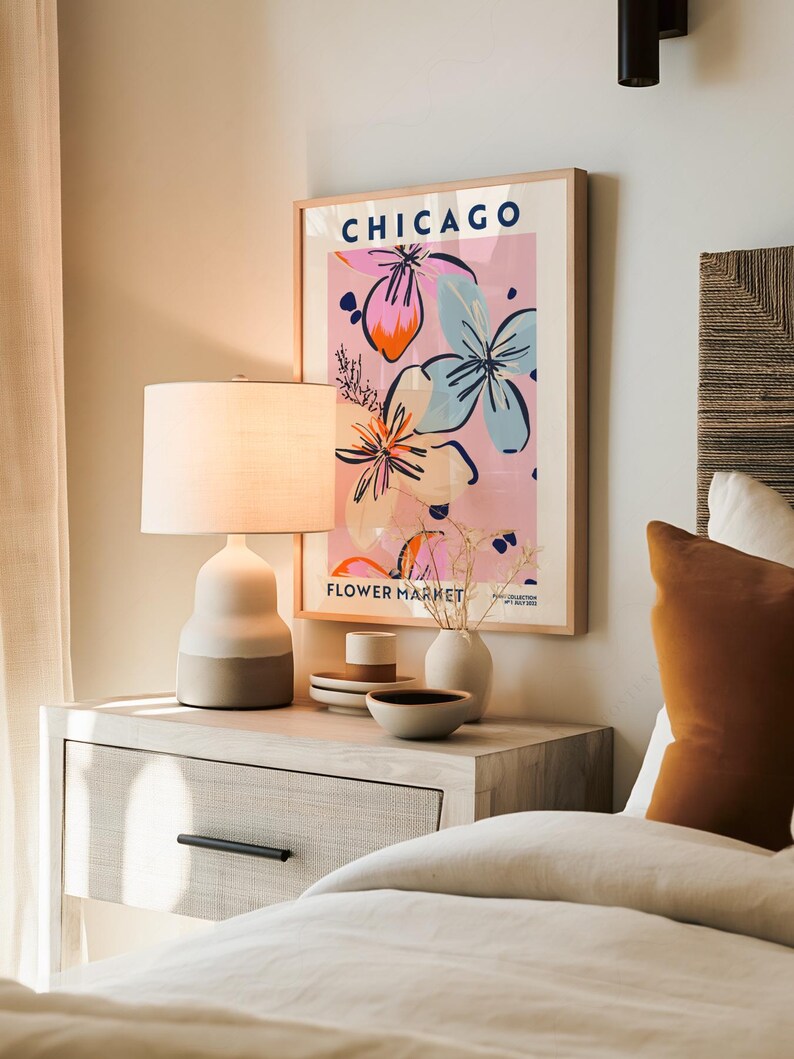 May include: A framed wall art print titled CHICAGO FLOWER MARKET with a pink background and abstract floral illustrations in blue, orange, and black. The print is displayed in a light wood frame above a bedside table with a lamp and ceramic bowls.
