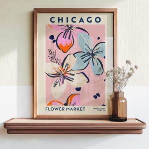 May include: A framed wall art print titled CHICAGO FLOWER MARKET with a pink background and abstract floral illustrations in shades of blue, orange, and cream. The text includes PRINT COLLECTION N&deg;1 JULY 2023 in the bottom right corner.