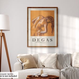 May include: A framed art print featuring a Degas drawing of a nude figure. The artwork is in warm tones and is displayed above a white sofa with pillows. The print has a brown frame and the artist's name is displayed in bold text.