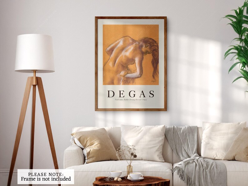 May include: A framed art print featuring a Degas drawing of a nude figure. The artwork is in warm tones and is displayed above a white sofa with pillows. The print has a brown frame and the artist's name is displayed in bold text.