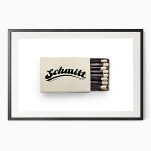 K&ouml;nnte beinhalten: Gerahmter Druck mit einer beigefarbenen Streichholzschachtel mit dem Namen "Schmitt" in Schwarz. Die Schachtel enth&auml;lt schwarze Streichh&ouml;lzer mit wei&szlig;en Spitzen. Das Bild ist vor einem wei&szlig;en Hintergrund.
