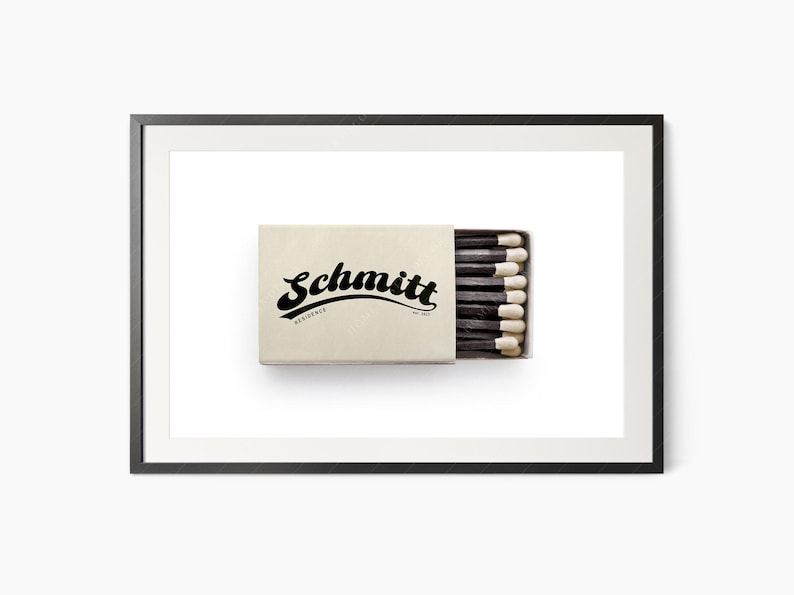 K&ouml;nnte beinhalten: Gerahmter Druck mit einer beigefarbenen Streichholzschachtel mit dem Namen "Schmitt" in Schwarz. Die Schachtel enth&auml;lt schwarze Streichh&ouml;lzer mit wei&szlig;en Spitzen. Das Bild ist vor einem wei&szlig;en Hintergrund.