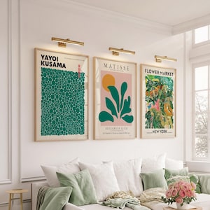 May include: Three framed art prints hanging on a white wall, each illuminated by a gold picture light. The left print features green dots by Yayoi Kusama, the center shows a Matisse design with a yellow sun, and the right depicts a floral market.
