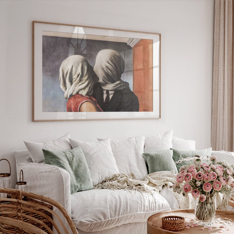 May include: A rectangular framed wall art print featuring a reproduction of The Lovers by Ren&eacute; Magritte, depicting two figures with their heads covered in white fabric, wearing a red top and a dark suit, displayed in a thin light wood frame.