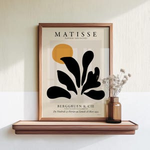 May include: A framed Matisse Papiers D&eacute;coup&eacute;s exhibition poster featuring a yellow circle and black organic leaf shapes on a beige background. The text reads MATISSE PAPIERS D&Eacute;COUP&Eacute;S BERGGRUEN & CIE 70 RUE DE L'UNIVERSIT&Eacute;, PARIS VII Du Vendredi 27 F&eacute;vrier au Samedi 28 Mars 1953.