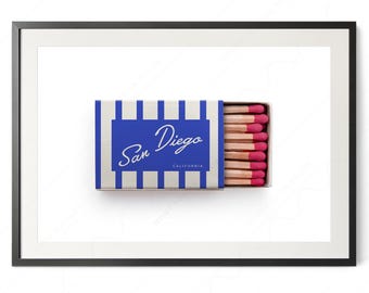 May include: A framed print featuring a matchbox with blue and white vertical stripes. The center blue rectangle displays the white cursive text San Diego and the word California in a smaller font. The box is partially open, revealing wooden matches.