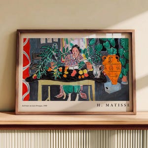May include: A framed art print of Henri Matisse's 1940 painting, Int&eacute;rieur au vase &eacute;trusque. The scene depicts a person sitting at a table with a black cloth, green plants, a large orange vase, and fruit. The text reads Int&eacute;rieur au vase &eacute;trusque, 1940 and H. MATISSE.