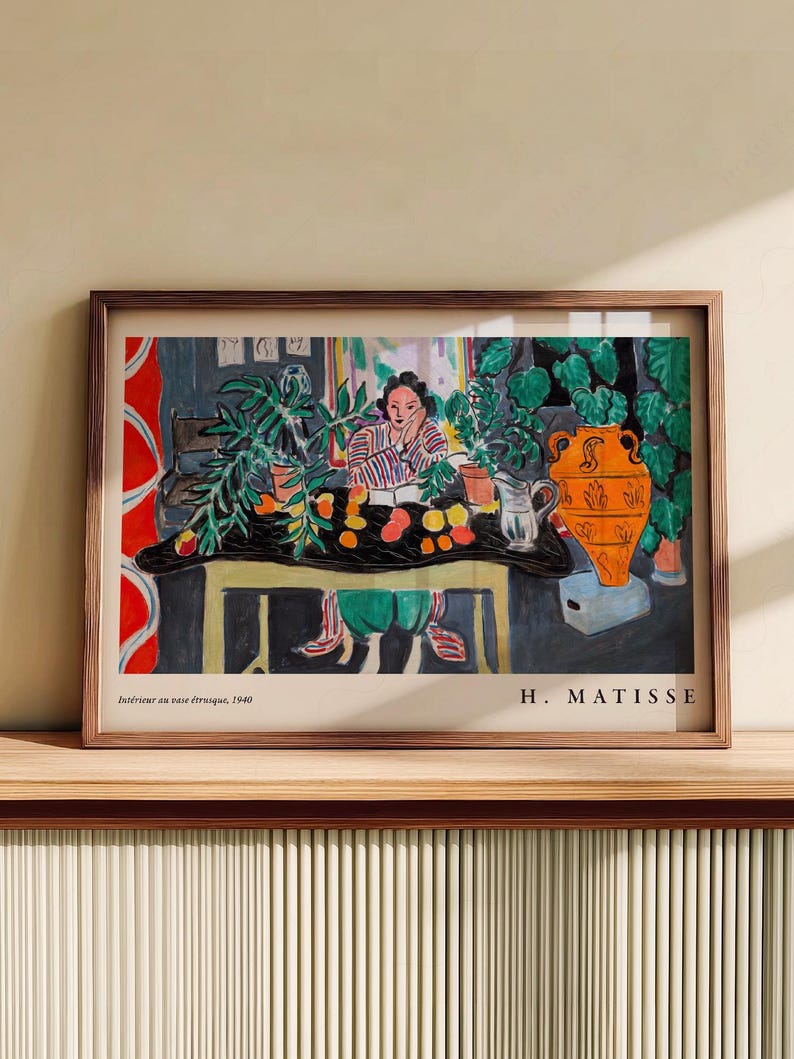 May include: A framed art print of Henri Matisse's 1940 painting, Int&eacute;rieur au vase &eacute;trusque. The scene depicts a person sitting at a table with a black cloth, green plants, a large orange vase, and fruit. The text reads Int&eacute;rieur au vase &eacute;trusque, 1940 and H. MATISSE.