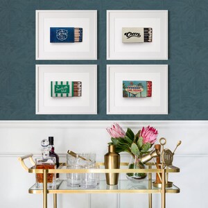 K&ouml;nnte beinhalten: Vier gerahmte Drucke von Streichholzschachteln mit verschiedenen Designs, darunter "Ritz", "Cheers", "The Beverly Hills Hotel" und eine Las Vegas-Szene. Ein goldener Servierwagen mit Glaswaren, Flaschen und Blumen befindet sich im Vordergrund.