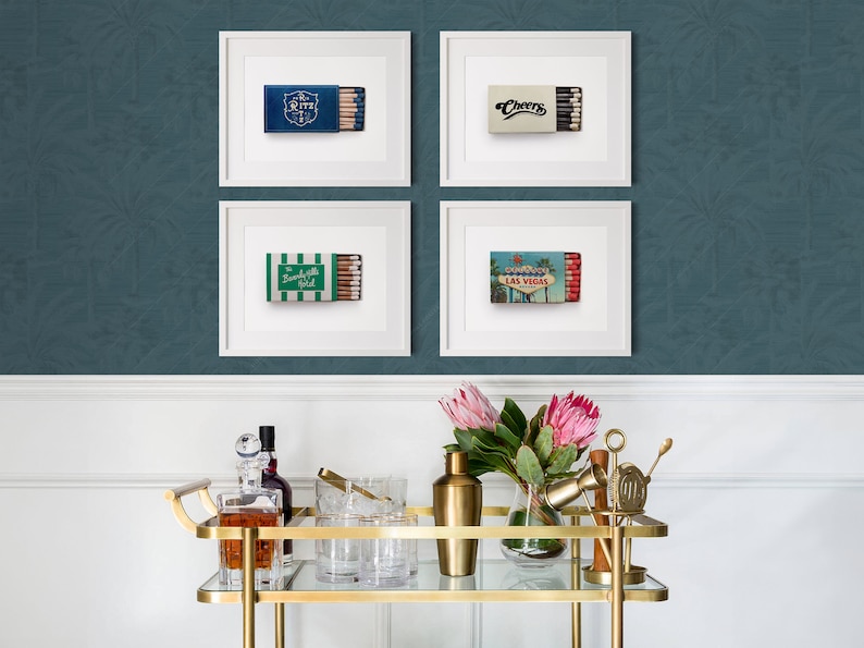 K&ouml;nnte beinhalten: Vier gerahmte Drucke von Streichholzschachteln mit verschiedenen Designs, darunter "Ritz", "Cheers", "The Beverly Hills Hotel" und eine Las Vegas-Szene. Ein goldener Servierwagen mit Glaswaren, Flaschen und Blumen befindet sich im Vordergrund.