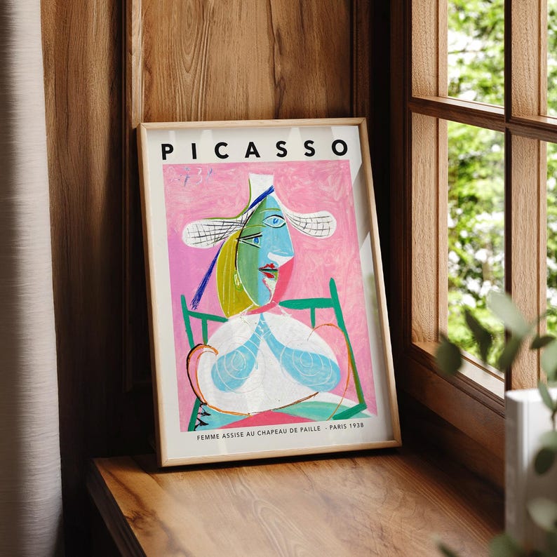 May include: A framed art print featuring a Picasso painting titled Femme Assise au Chapeau de Paille, Paris 1938. The artwork depicts a stylized figure in a straw hat with blue, green, and white tones against a vibrant pink background.