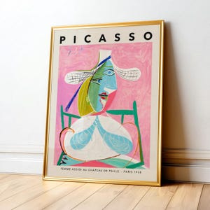 May include: A framed art print featuring Pablo Picasso's painting titled Femme Assise au Chapeau de Paille, Paris 1938. The artwork displays a stylized figure in a straw hat against a pink background, set within a thin, metallic gold frame.