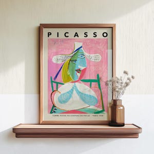 May include: A framed art print titled PICASSO FEMME ASSISE AU CHAPEAU DE PAILLE PARIS 1938 featuring a cubist portrait of a figure in a straw hat against a pink background, displayed on a wooden shelf next to a glass vase with dried flowers.