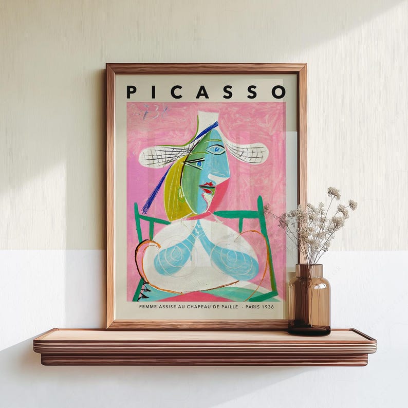 May include: A framed art print titled PICASSO FEMME ASSISE AU CHAPEAU DE PAILLE PARIS 1938 featuring a cubist portrait of a figure in a straw hat against a pink background, displayed on a wooden shelf next to a glass vase with dried flowers.