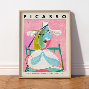 May include: A framed art print featuring a Picasso painting titled Femme Assise au Chapeau de Paille, Paris 1938. The artwork depicts a stylized figure in a wide-brimmed hat, rendered in shades of blue, green, yellow, and pink on a textured pink background.