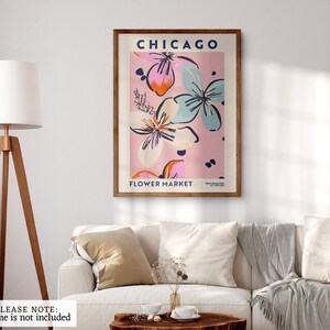 May include: A rectangular wall art print featuring the text CHICAGO FLOWER MARKET in dark blue. The design displays stylized floral illustrations in shades of pink, light blue, and orange against a soft pink background, presented in a wooden frame.