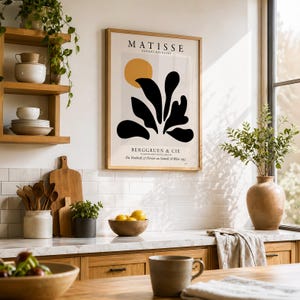 May include: A framed Matisse Papiers D&eacute;coup&eacute;s exhibition poster featuring a yellow circle and black organic shapes on a light background, hanging on a wall above a kitchen counter with wooden shelves, a bowl of lemons, and a ceramic vase.