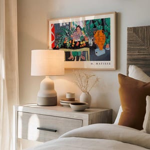 May include: A framed H. Matisse art print featuring a colorful interior scene with a person, plants, and an orange vase. The print is displayed above a light-colored nightstand with a two-toned table lamp, a small vase with dried stems, and bowls.