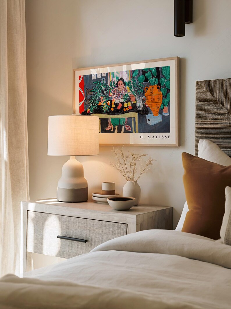 May include: A framed H. Matisse art print featuring a colorful interior scene with a person, plants, and an orange vase. The print is displayed above a light-colored nightstand with a two-toned table lamp, a small vase with dried stems, and bowls.