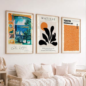 May include: Three framed art prints hanging on a wall. The left print shows a colourful coastal scene with the text Côte d'Azur. The middle print features black abstract shapes and an orange circle with the text Matisse. The right print displays a field of orange dots with the text Tokyo Modern Art.