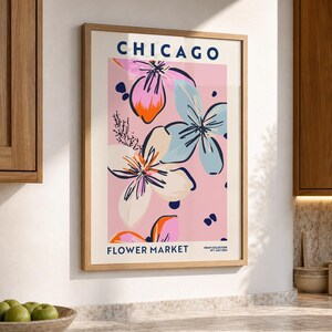 May include: A framed wall art print featuring the text CHICAGO FLOWER MARKET and PRINT COLLECTION N&deg;1 JULY 2022. The graphic design displays stylized flowers in shades of pink, light blue, orange, and dark blue against a soft pink background.