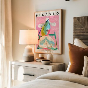 May include: A framed Picasso print titled Femme Assise au Chateau de Paris 1938 featuring a colorful abstract portrait on a pink background, displayed above a bedside table with a two-toned ceramic lamp, a small vase, and stacked bowls.