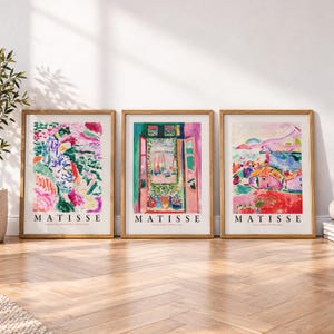 May include: Three framed art prints featuring colorful Matisse paintings. Each print displays a distinct landscape or interior scene in vibrant hues of pink, green, blue, and red, with the name MATISSE printed in black capital letters at the bottom.