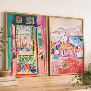 May include: Two rectangular framed art prints featuring vibrant, impressionistic paintings. The left print depicts an open window overlooking a harbor with sailboats. The right print shows a coastal town with pink buildings and blue water.