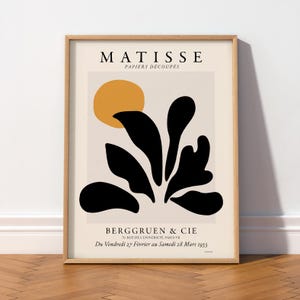 May include: A framed art print featuring the text MATISSE PAPIERS D&Eacute;COUP&Eacute;S at the top and BERGGRUEN & CIE at the bottom. The design includes a solid yellow circle and abstract black organic shapes on a light beige background in a wooden frame.