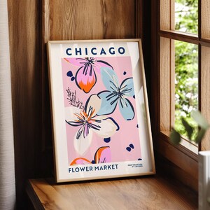 May include: A framed vertical art print featuring the text CHICAGO FLOWER MARKET and PRINT COLLECTION NO 1 JULY 2022. The design displays stylized floral illustrations in shades of light blue, orange, and white against a soft pink background.