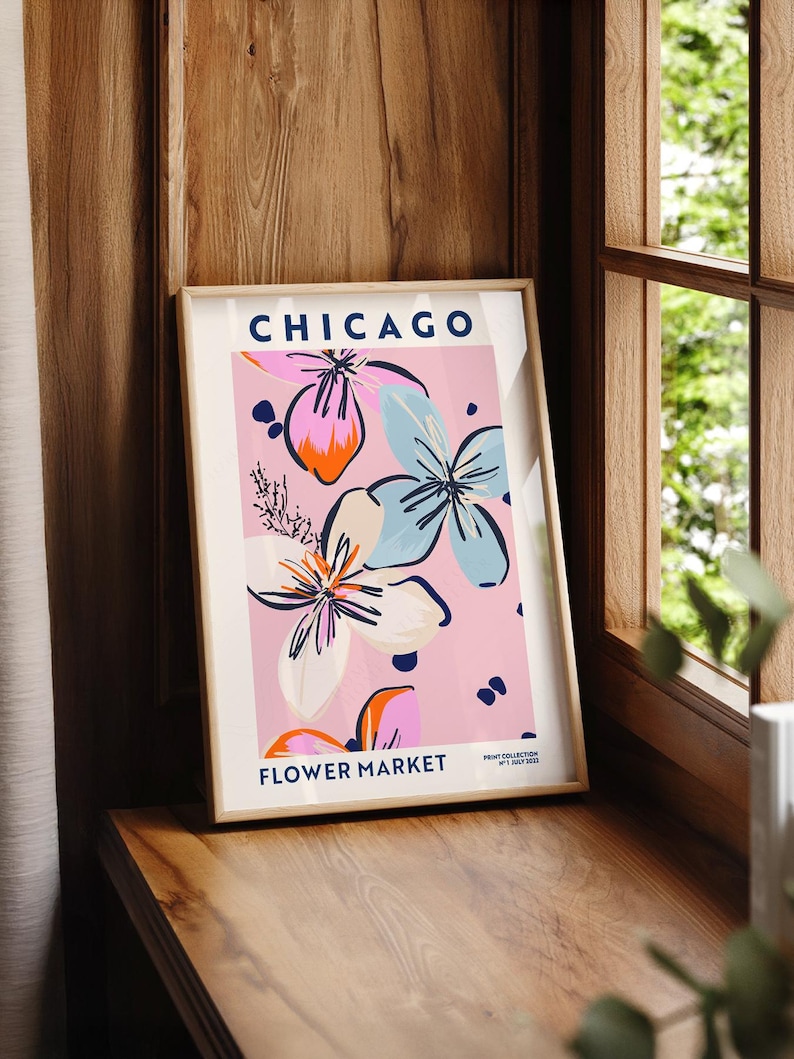 May include: A framed vertical art print featuring the text CHICAGO FLOWER MARKET and PRINT COLLECTION NO 1 JULY 2022. The design displays stylized floral illustrations in shades of light blue, orange, and white against a soft pink background.