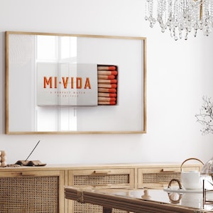 K&ouml;nnte beinhalten: Gerahmtes Kunstwerk mit einem Streichholzschachtel-Design, den Worten "MI VIDA" in Orange. Die Streichholzschachtel ist ge&ouml;ffnet und zeigt Streichh&ouml;lzer mit roten Spitzen. Das Kunstwerk h&auml;ngt &uuml;ber einem Holzschrank.