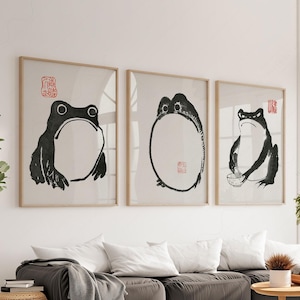 May include: Three framed art prints with minimalist black and white frog illustrations. Each print shows a different frog pose, with a red stamp in the upper corner. The frames are a light wood colour.