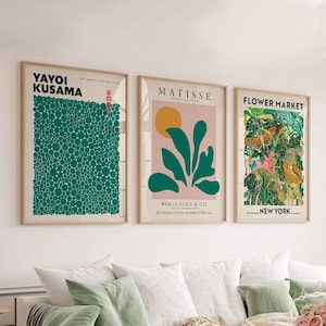 May include: Three framed art prints hanging on a wall. The left print features Yayoi Kusama text and a green dot pattern. The center print shows Matisse text, a yellow sun, and green leaves on a pink background. The right print displays Flower Market New York text with a colorful botanical illustration.