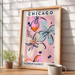 May include: A framed vertical art print featuring the text CHICAGO FLOWER MARKET in dark blue. The design displays stylized floral illustrations in shades of pink, light blue, orange, and dark blue against a soft pink background.