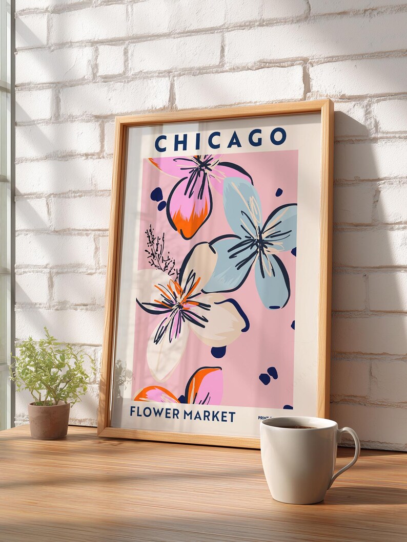 May include: A framed vertical art print featuring the text CHICAGO FLOWER MARKET in dark blue. The design displays stylized floral illustrations in shades of pink, light blue, orange, and dark blue against a soft pink background.