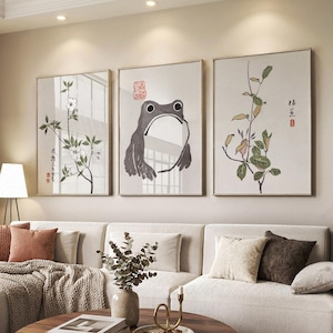 May include: A set of three rectangular wall art prints featuring minimalist ink-style illustrations. The left print shows white flowers on a branch, the centre print depicts a stylised grey frog, and the right print displays a green leafy plant.