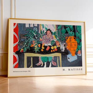 May include: A framed art print of Henri Matisse's 1940 painting Int&eacute;rieur au vase &eacute;trusque, featuring a person at a table with fruit, plants, and an orange vase. The print has a gold frame and includes the artist's name and the title at the bottom.