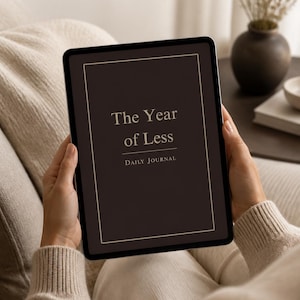 May include: A digital tablet displaying a dark brown cover with a thin cream-colored border. The text on the screen reads The Year of Less in a serif font, followed by the words DAILY JOURNAL in a smaller, capitalized sans-serif typeface.