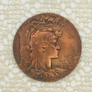 Op de afbeelding: Een bronzen medaille met een profiel van een vrouw die een krans van bladeren draagt en een afbeelding van een stadsbeeld. De medaille is gegraveerd met "REPUBLIQUE FRANCAISE".