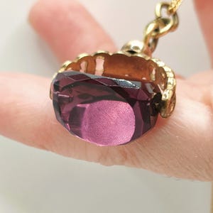 Può includere: Una collana con catena d'oro con un ciondolo in vetro viola. Il ciondolo è incastonato in una cornice dorata con un design decorativo.