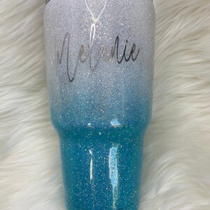 May include: A white and blue ombre glitter tumbler with the name "Melanie" written in silver script. A small silver balloon graphic is above the name.