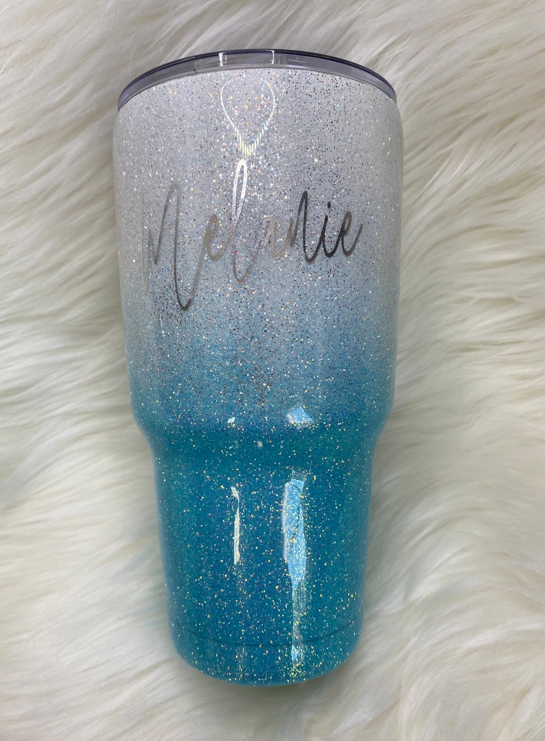 May include: A white and blue ombre glitter tumbler with the name "Melanie" written in silver script. A small silver balloon graphic is above the name.